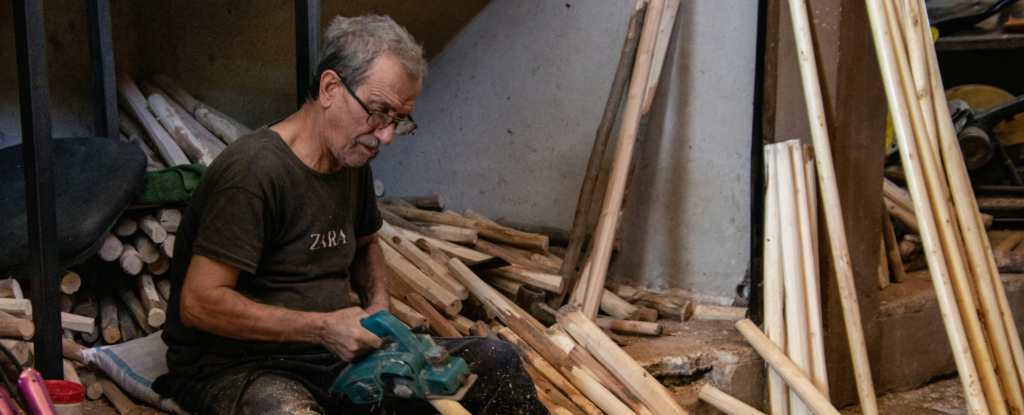A man uses a handheld power tool to smooth a wooden dowel while sitting barefoot in a workshop filled with wooden sticks.