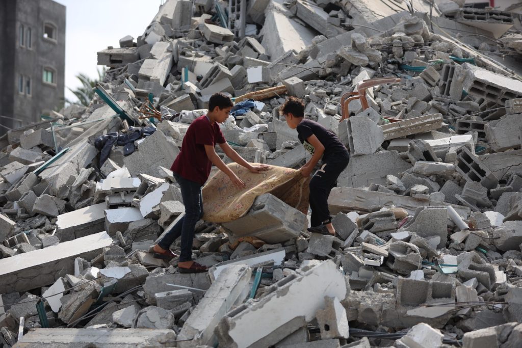 Two boys search through rubble, carrying a large cushion amidst debris from collapsed concrete buildings in Gaza, Occupied Palestinian Territories.