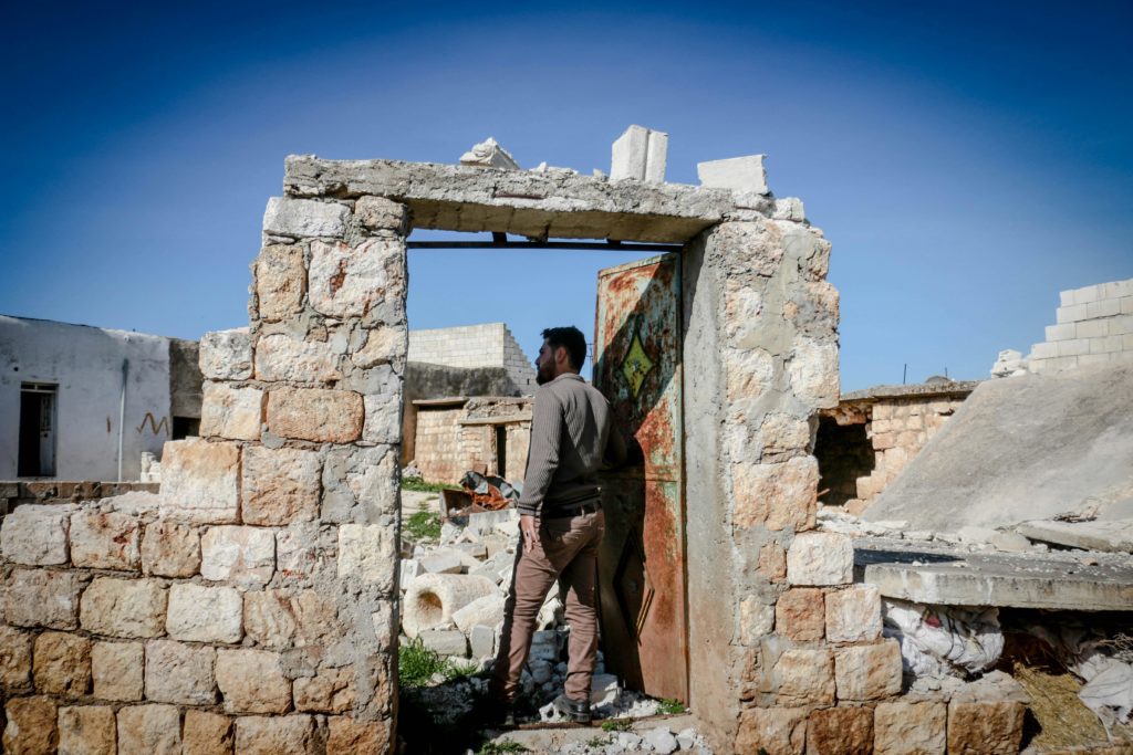 A man stands in a doorway of a crumbling brick structure on a clear day, surrounded by rubble and remnants of buildings in Idlib, Syria.