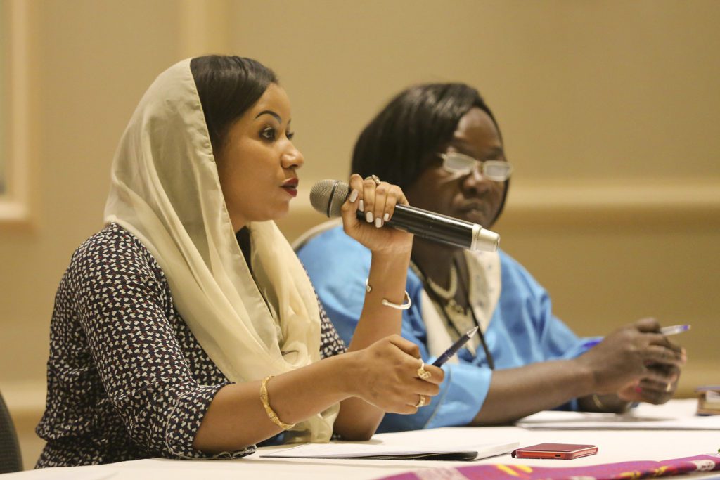 Two black women sit at a table during a formal meeting. One speaks into a microphone while the other listens attentively with a pen in hand.