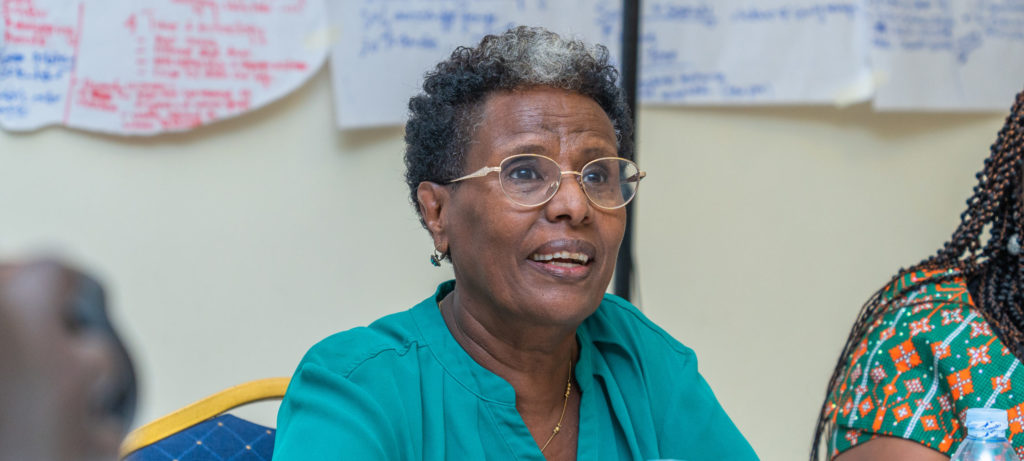 Elderly black woman in a green shirt speaks during a discussion meeting with notes and water bottles on the table.