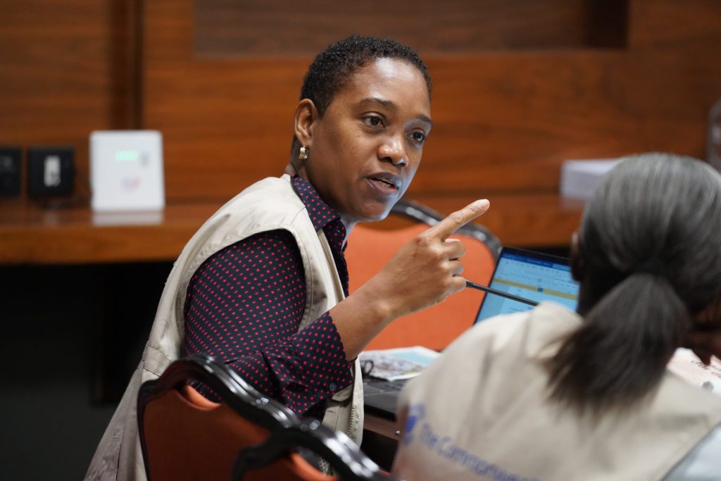 A black woman with short hair wearing a beige vest gestures while talking to another person at a conference, with a laptop and wood-panel wall in the background.